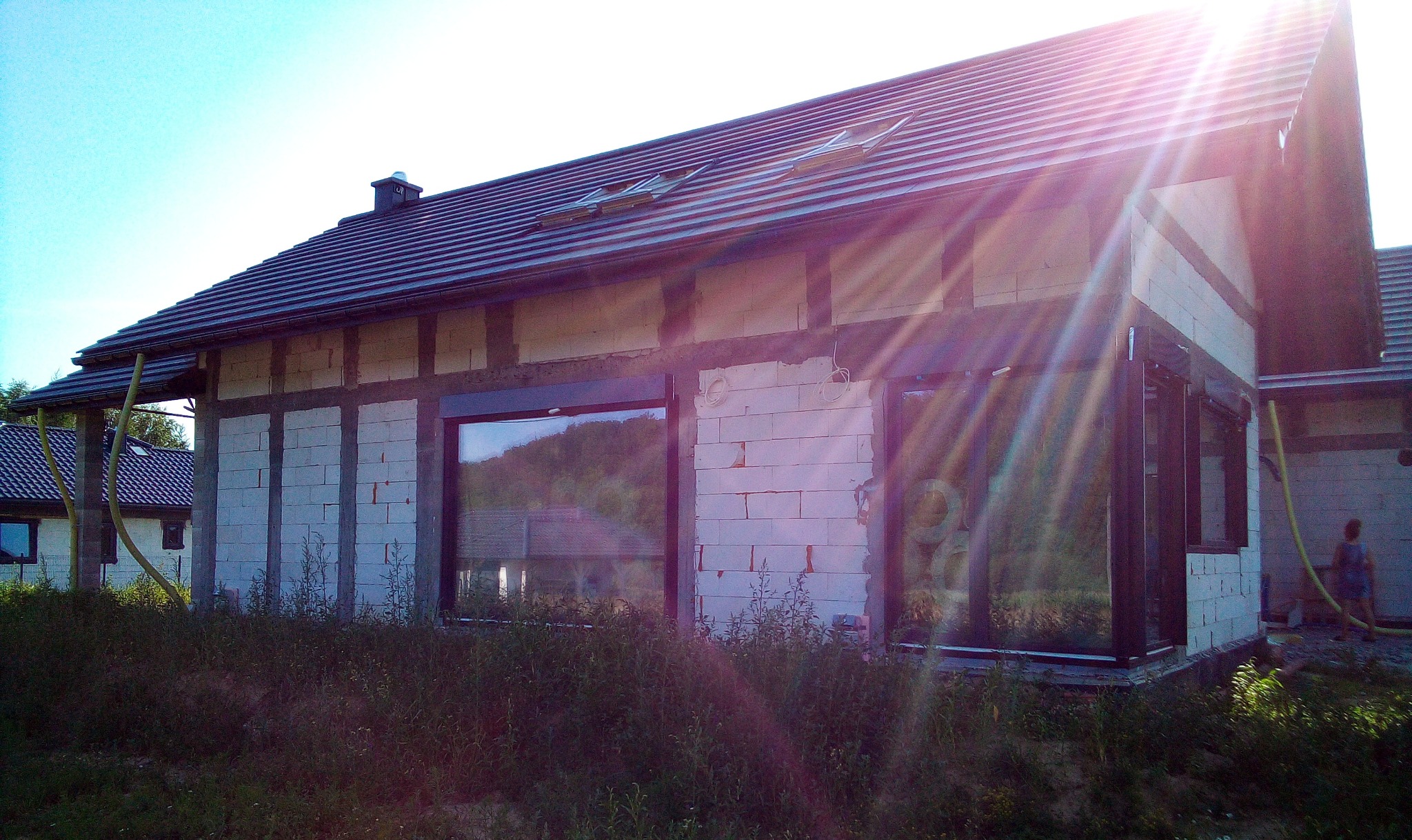 Surowy budynek w trakcie budowy z zamontowanymi oknami w czarnych ramach, widok z zewnątrz na tle wysokiej trawy, oświetlony silnym słońcem.