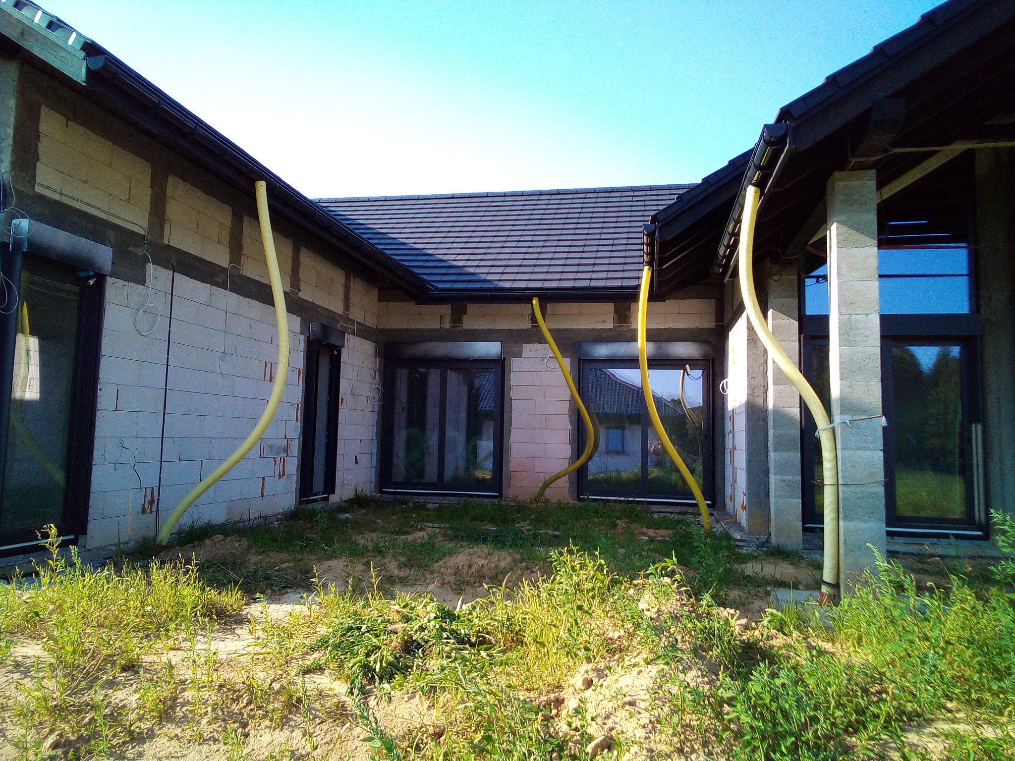 Surowy stan budynku w trakcie budowy z zamontowanymi oknami z czarnymi ramami i żółtymi rurami spustowymi przymocowanymi do elewacji, widoczne bloczki betonowe i niezagospodarowany teren.