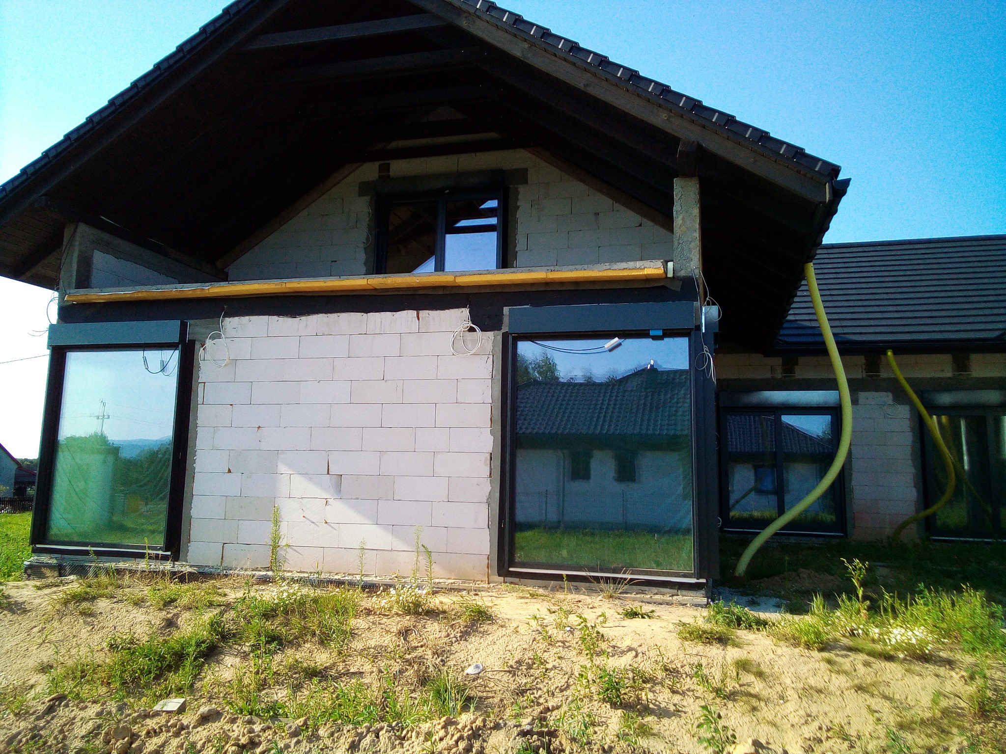 Budynek w trakcie budowy z zamontowanymi oknami PCV z czarnymi ramami, widoczne przewody instalacyjne i surowy stan elewacji.