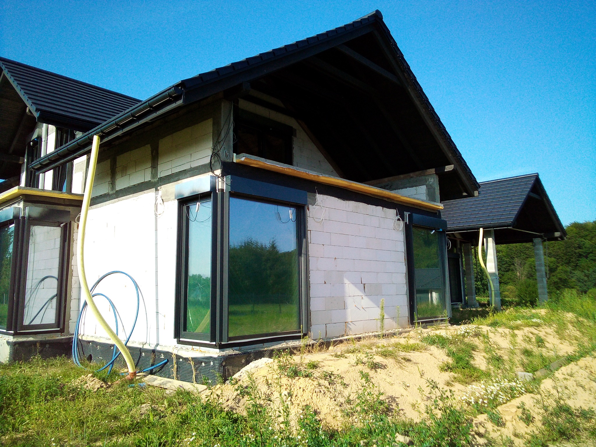 Surowy stan budynku z czarnymi oknami i instalacją zewnętrzną na tle zieleni. Widoczny etap montażu stolarki okiennej w nowym domu.