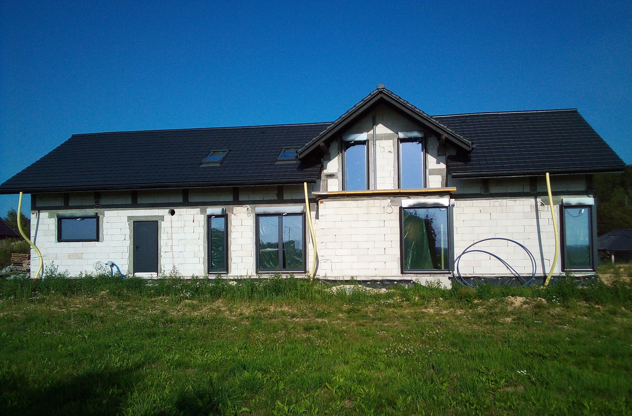 Surowy stan domu jednorodzinnego z zamontowanymi oknami w czarnych ramach, widoczne rury i folie ochronne na oknach, trawa na pierwszym planie.