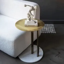 Złoty stolik pomocniczy z marmurową podstawą, na którym stoi rzeźba dyskobola na książce 'Michelangelo', obok biały, teksturowany fotel.
