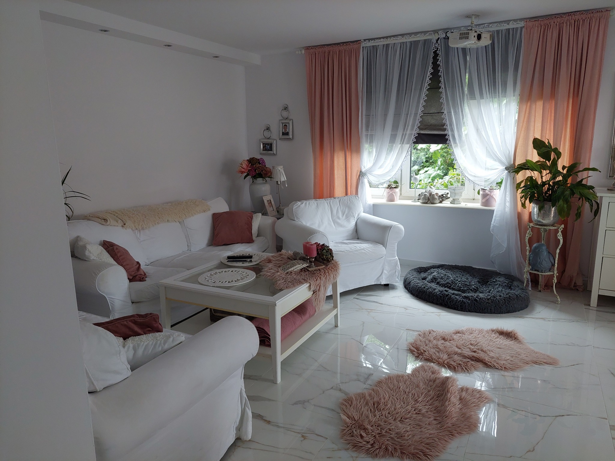 Elegancki salon w Łubowie z białymi sofami, marmurową podłogą i dekoracyjnymi zasłonami. Widoczny projektor sufitowy i rośliny doniczkowe.