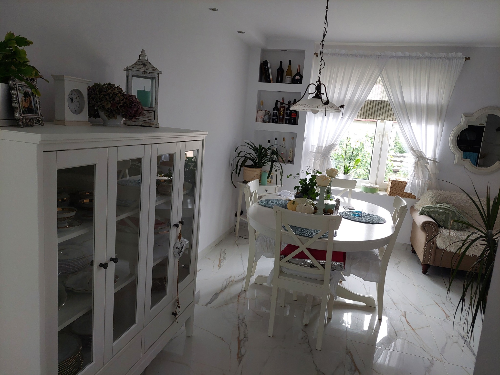 Jasne wnętrze jadalni z białymi meblami: kredens z przeszkleniami, okrągły stół z dekoracjami, białe krzesła i marmurowe płytki podłogowe z delikatnym żyłowaniem.