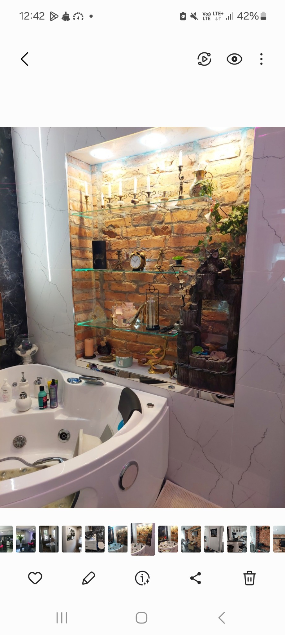 Nowoczesna łazienka z narożną wanną jacuzzi i dekoracyjną wnęką z cegły, podświetlonymi szklanymi półkami, świecznikami, zegarem i kaskadową fontanną.