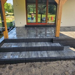 TOMASZ BURZA - Nowoczesne schody wejściowe z kostki brukowej w odcieniach szarości i czerni, prowadzące do domu z drewnianą konstrukcją werandy. W tle widoczne odbicie osoby w oknie.