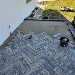 TOMASZ BURZA - Układanie kostki brukowej w jodełkę na podsypce piaskowej. Widoczny gumowy młotek, wiadro z narzędziami i wytyczone sznurki. Częściowo ułożona nawierzchnia.