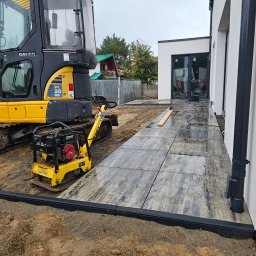 TOMASZ BURZA - Układanie kostki brukowej na posesji z użyciem zagęszczarki i minikoparki. Widoczne świeżo ułożone płyty chodnikowe i przygotowane podłoże.