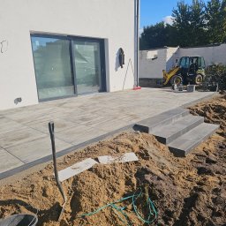 TOMASZ BURZA - Układanie kostki brukowej na tarasie z widocznymi schodami, koparką w tle i narzędziami. Etap prac ziemnych, przygotowanie podłoża. Jasne płytki tarasowe.