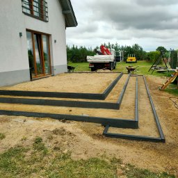 TOMASZ BURZA - Przygotowanie podłoża pod taras z obrzeżami w Wolinie. Widoczny piasek, obrzeża z tworzywa, częściowo trawa. W tle dom i samochód dostawczy z dźwigiem.