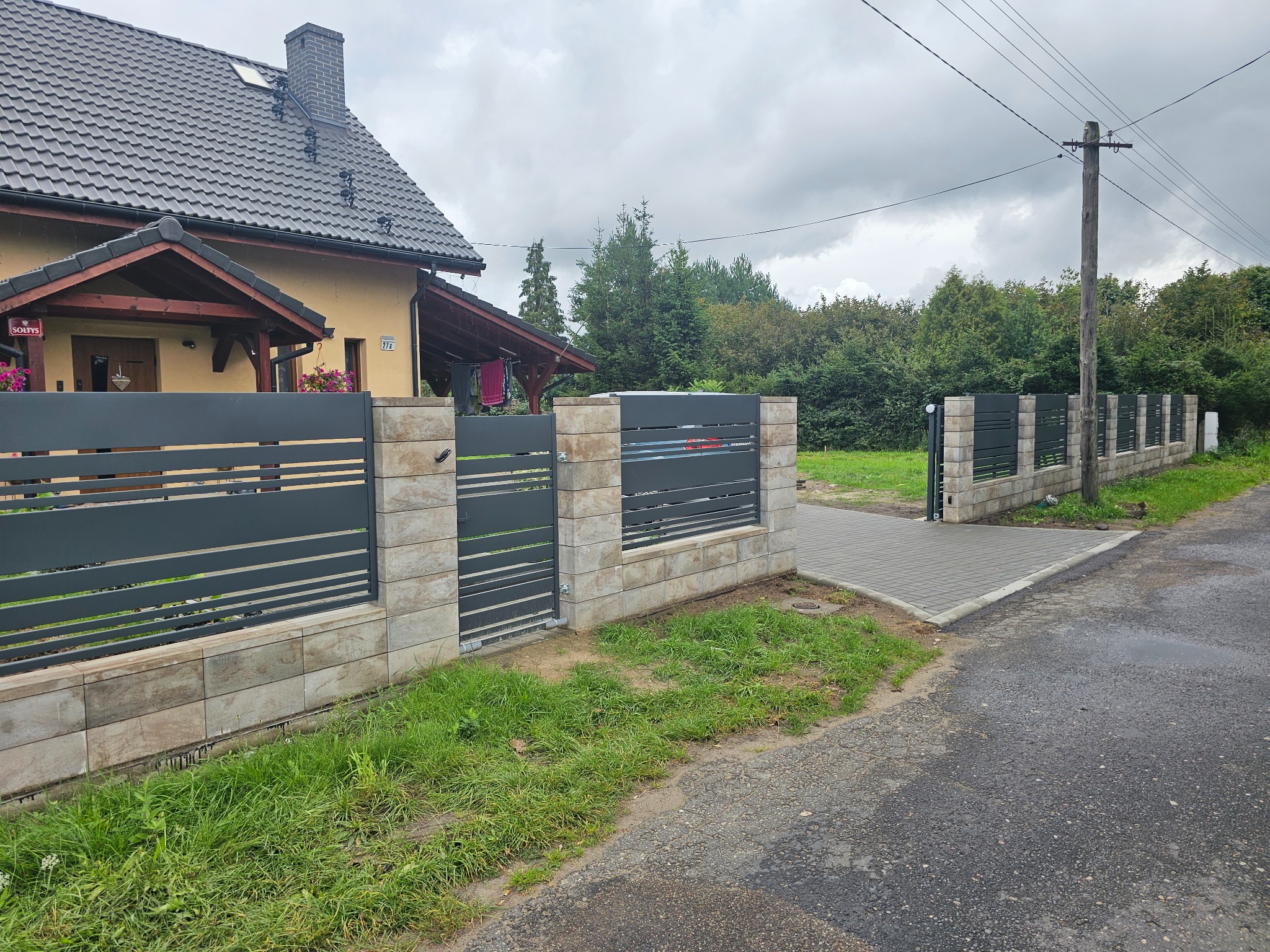 Nowoczesne ogrodzenie posesji z szarego metalu i kamiennych słupków, z bramą wjazdową i furtką, na tle domu z ciemną dachówką i zieleni. Widok z ulicy.