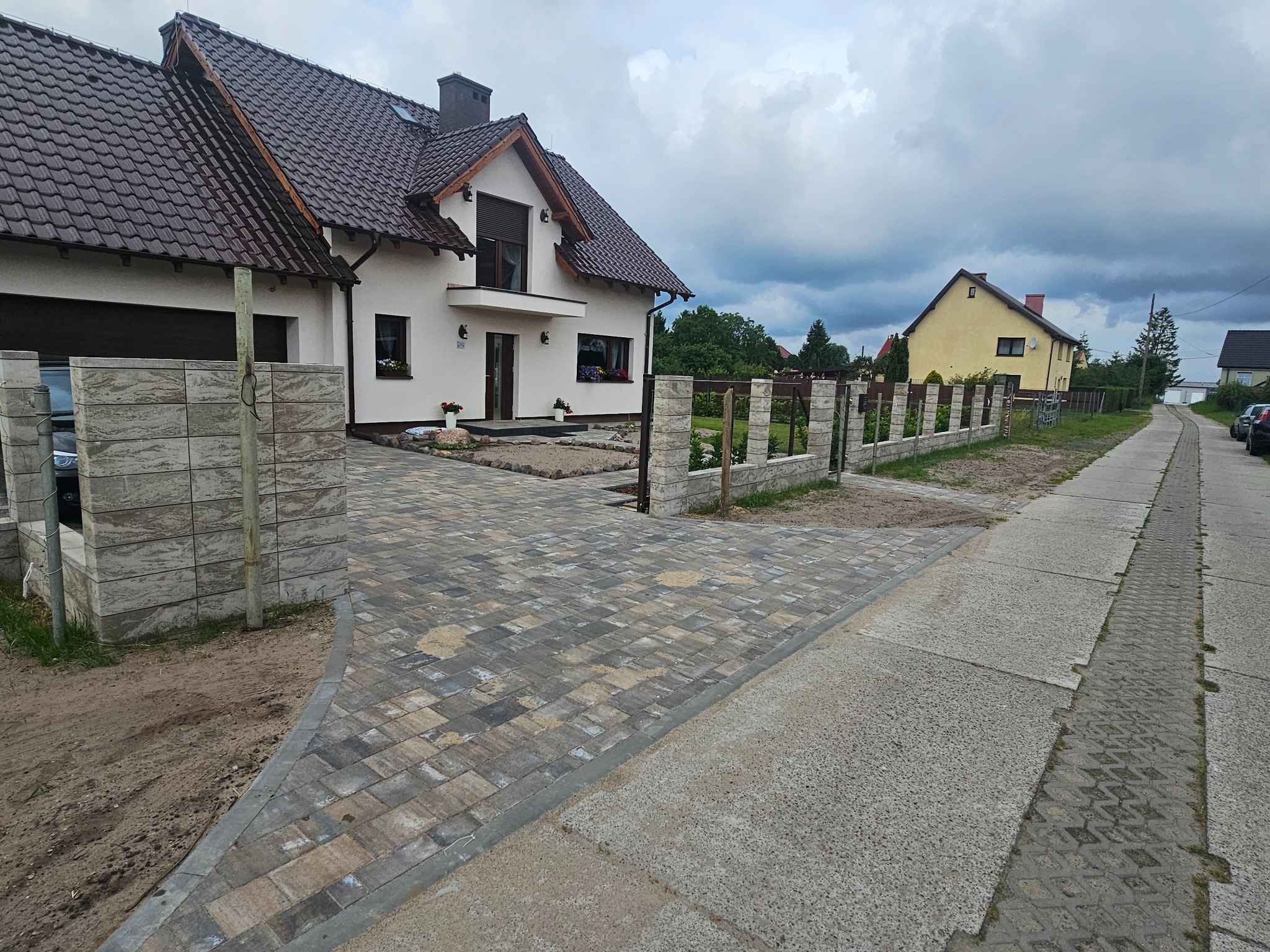 Nowoczesny dom jednorodzinny z brązowym dachem i jasną elewacją, otoczony kostką brukową w odcieniach szarości. Widok z poziomu ulicy, pochmurne niebo w tle.