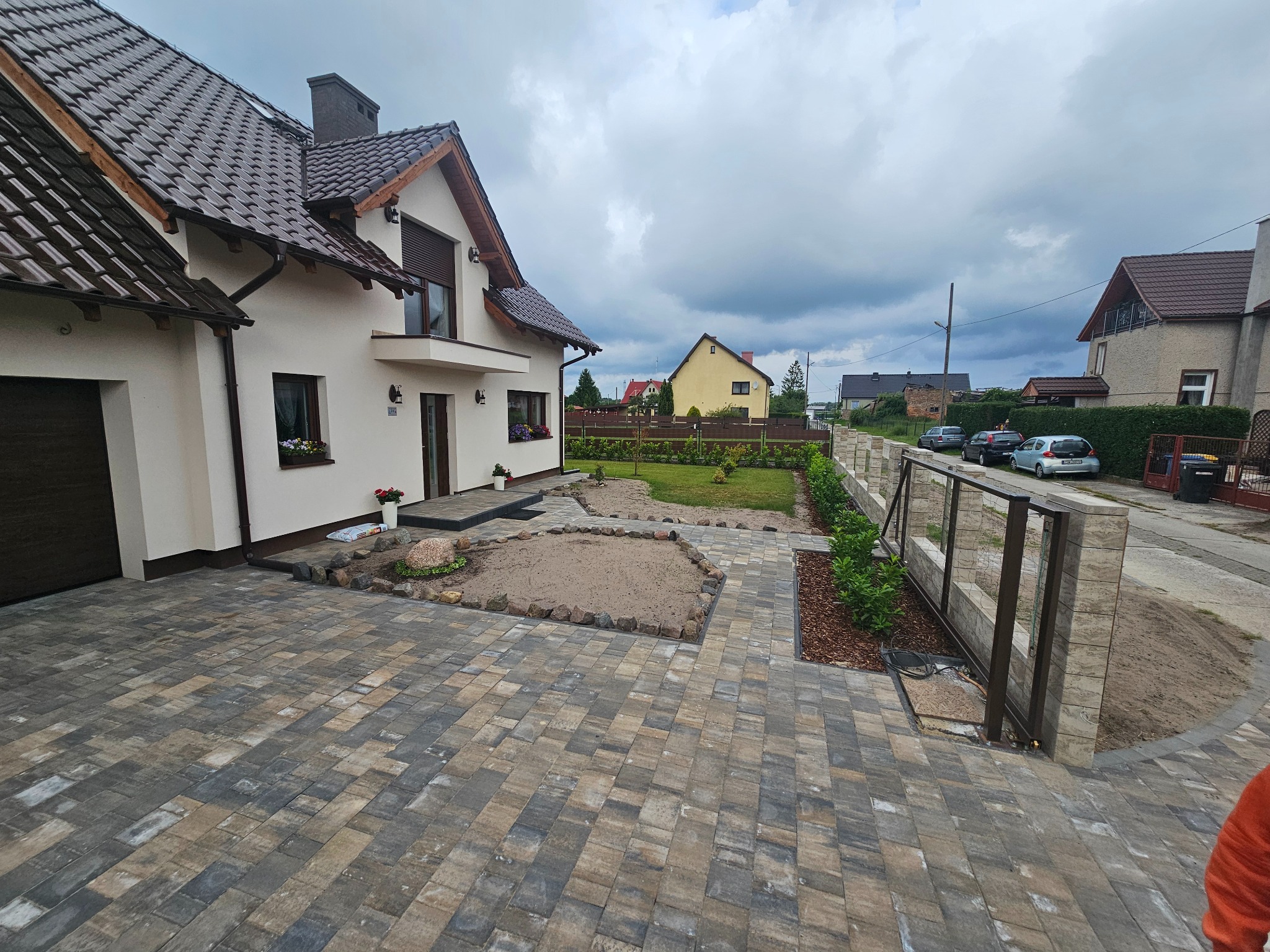 Ułożona kostka brukowa na podjeździe przed domem z ogrodem. Widoczne elementy architektury krajobrazu i ogrodzenia. Dom z brązowym dachem i białymi ścianami.