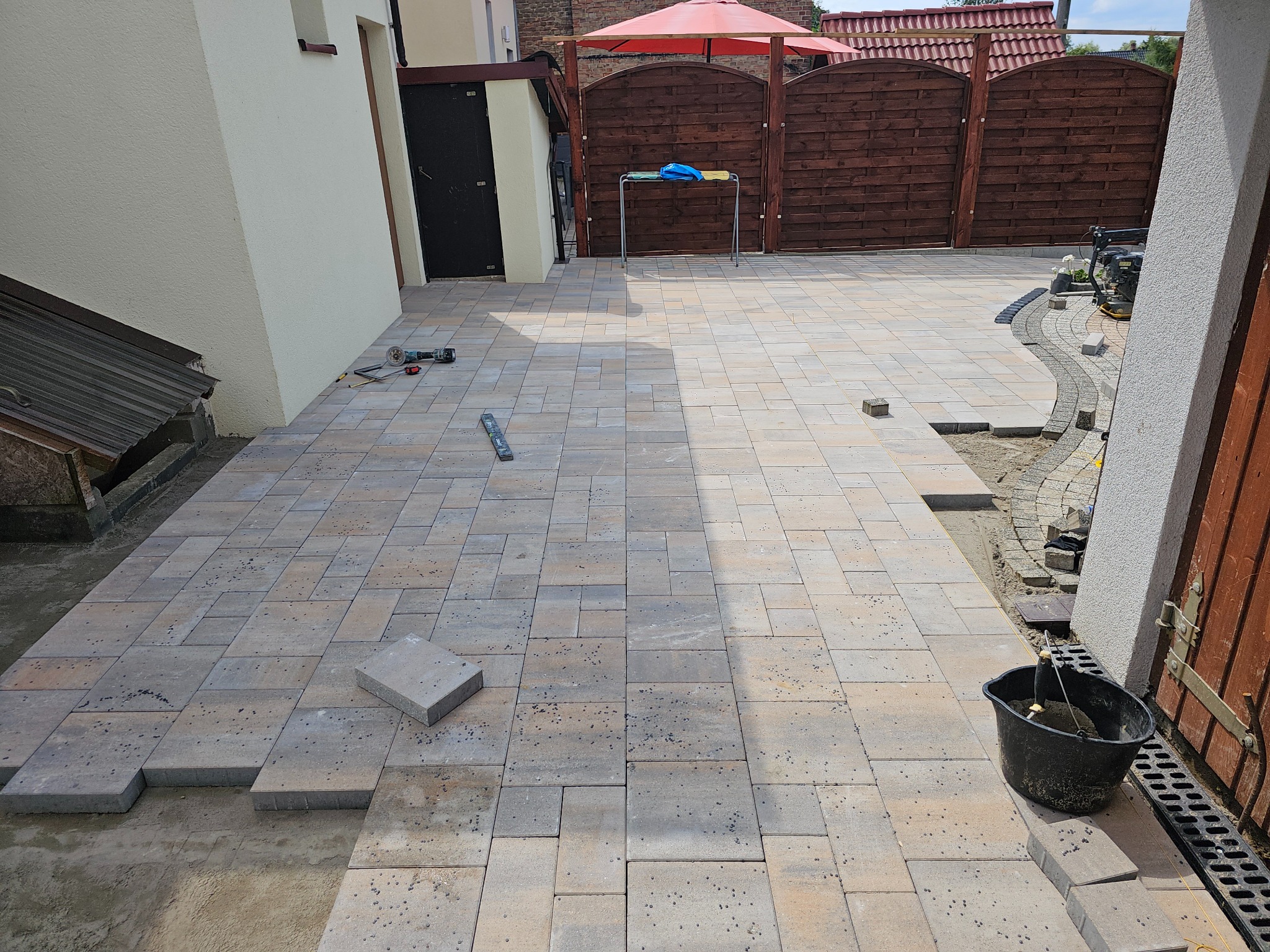 Układanie kostki brukowej na posesji, widoczne narzędzia i częściowo ułożona nawierzchnia. Prace brukarskie w toku, zarys ogrodu i elementy architektury.