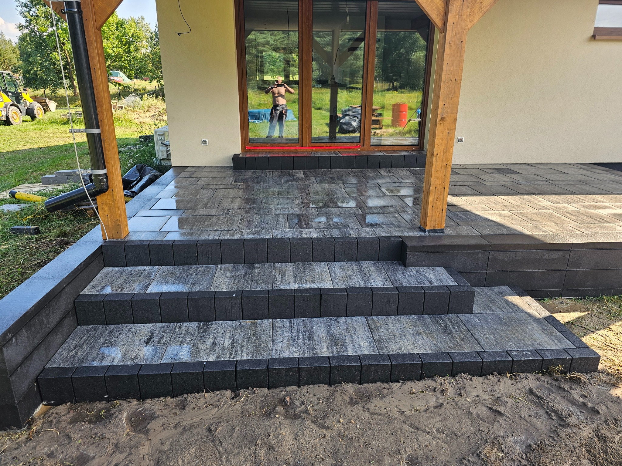 Nowoczesne schody wejściowe z kostki brukowej w odcieniach szarości i czerni, prowadzące do domu z drewnianą konstrukcją werandy. W tle widoczne odbicie osoby w oknie.