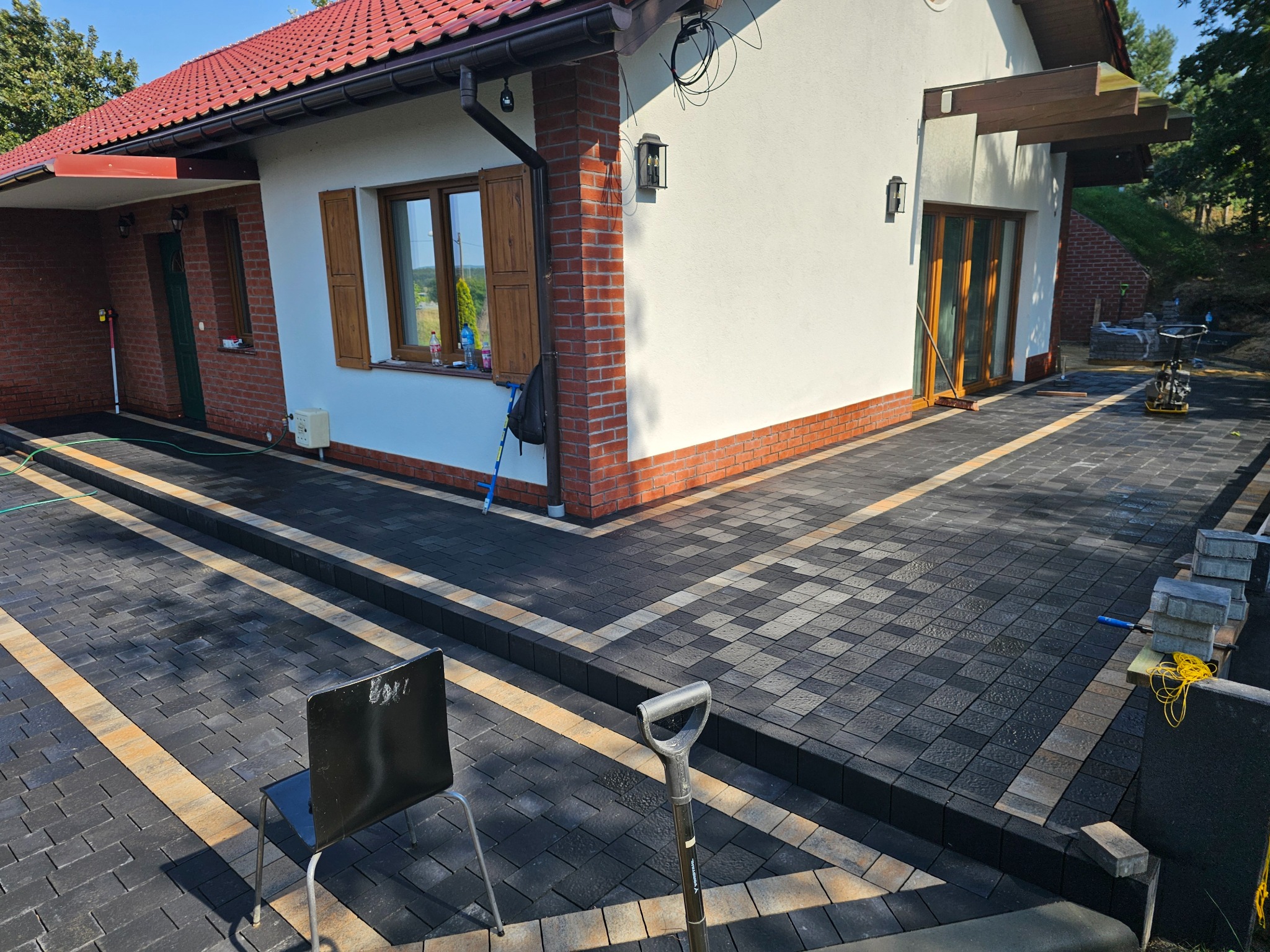 Ułożona kostka brukowa w odcieniach szarości i beżu przed domem z czerwoną dachówką. Widoczny fragment elewacji i narzędzia brukarskie.