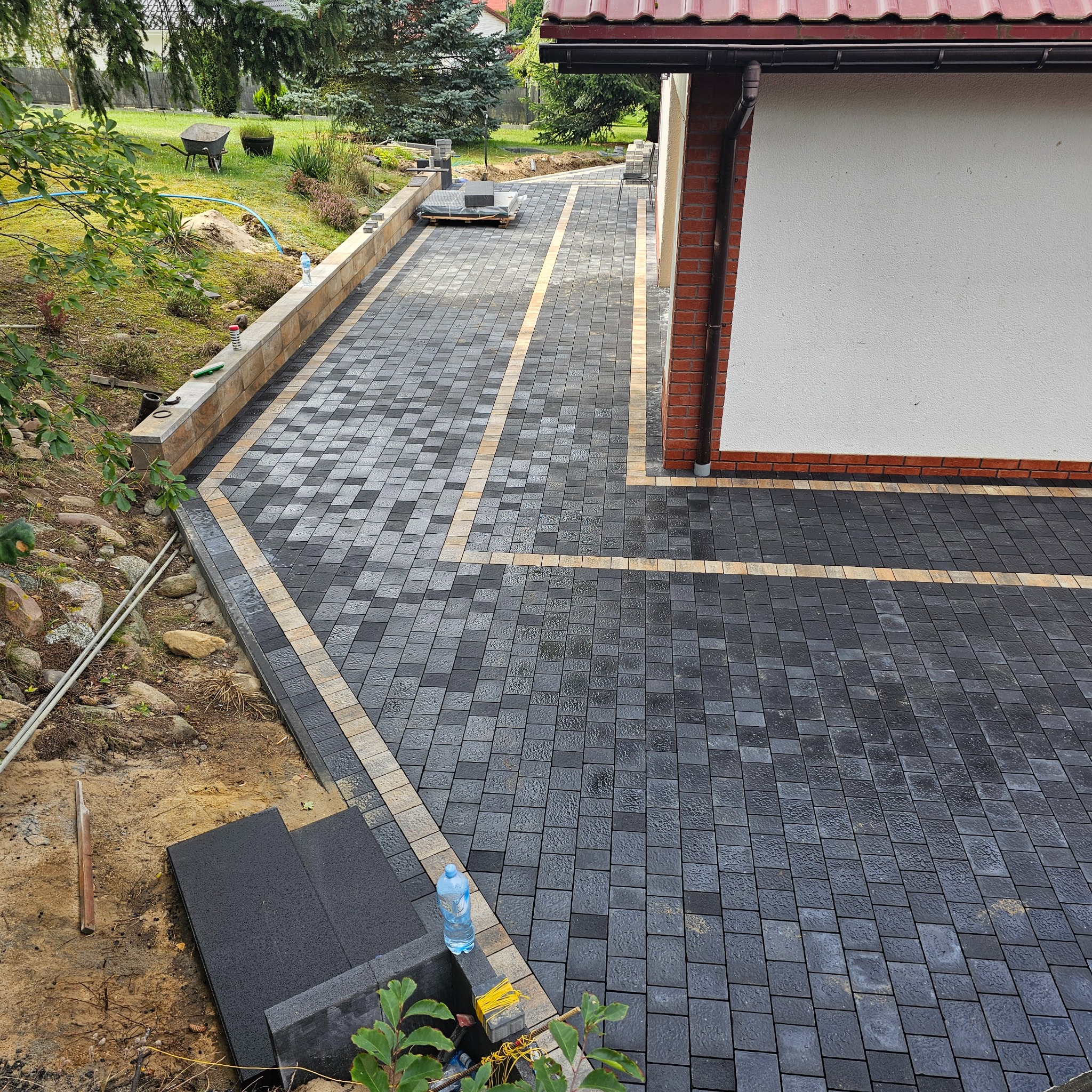 Ułożona kostka brukowa w odcieniach szarości i beżu, tworząca wzór na podjeździe. Widoczne obrzeża i fragment budynku z ceglaną elewacją. Częściowo widoczny trawnik.