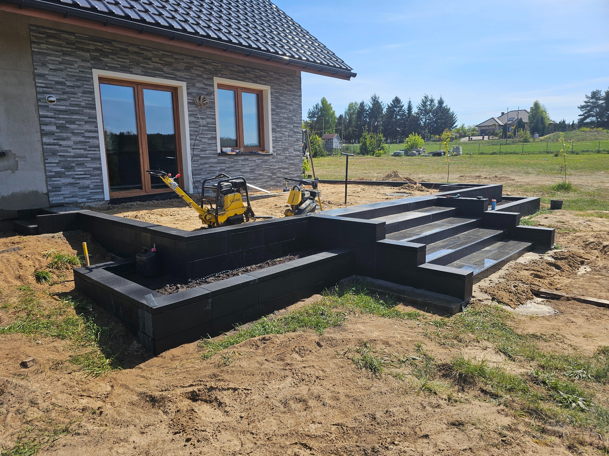 Nowoczesny taras z czarnych płyt, w trakcie budowy. Widoczne zagęszczarki i piasek. Dom z szarą elewacją w tle. Słoneczny dzień.