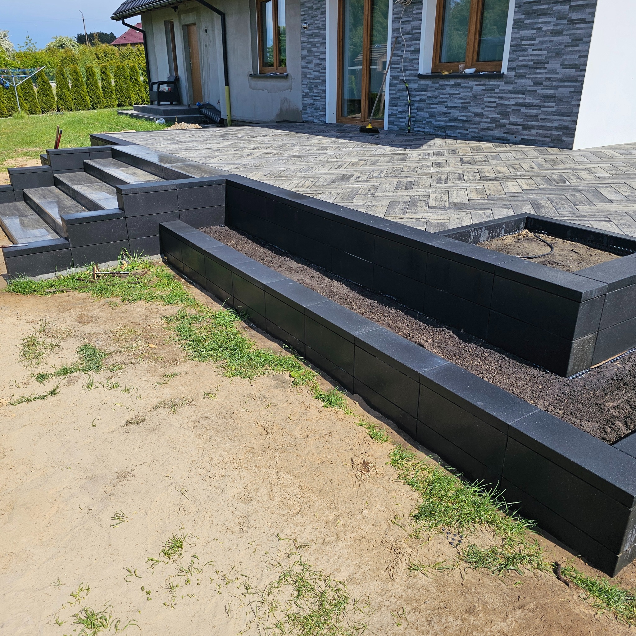 Nowoczesny taras z ciemnej kostki brukowej, schody i podwyższone rabaty z czarnych bloczków. Widok na dom z szarą elewacją i drewnianymi oknami. Częściowo zagospodarowany teren.
