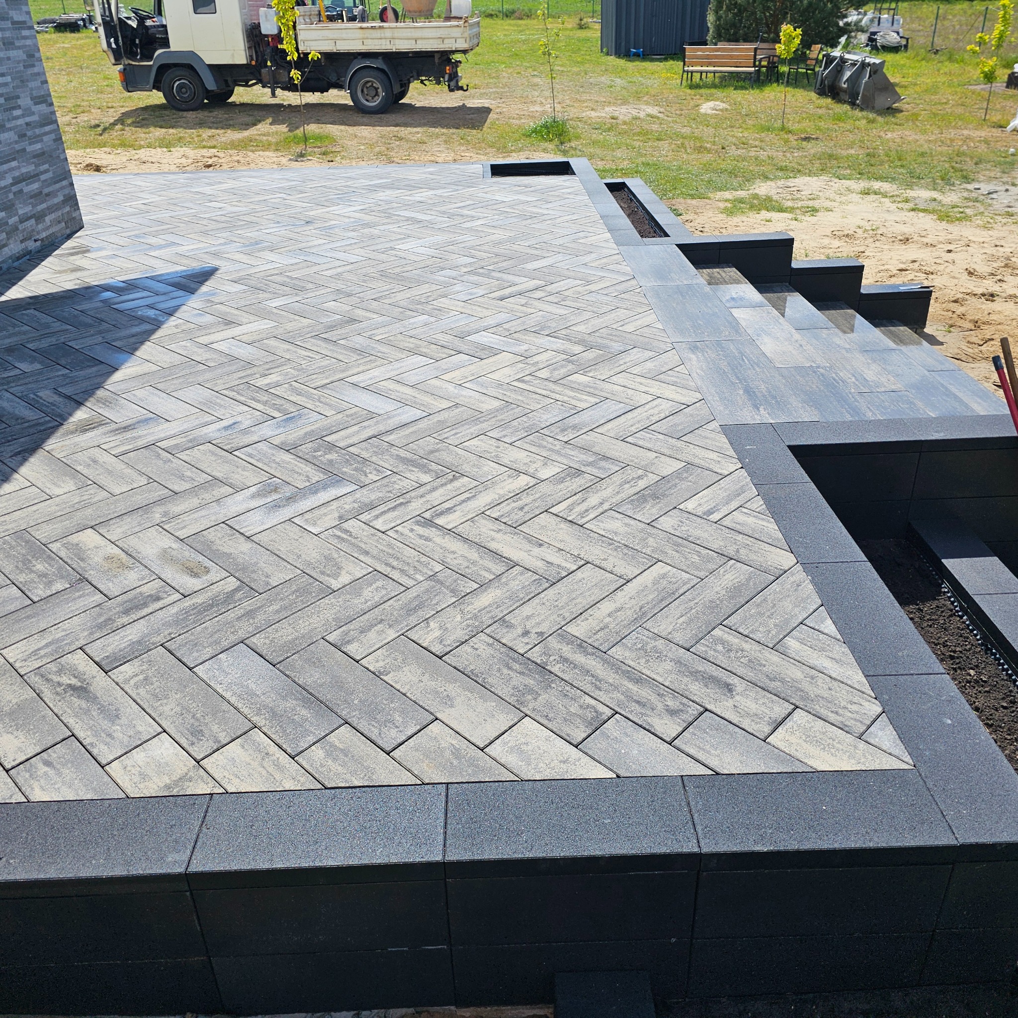 Ukończony taras z szarej kostki ułożonej w jodełkę, obramowany czarnym obrzeżem. Widoczne schody i fragment ogrodu z pojazdem w tle. Całość w słoneczny dzień.