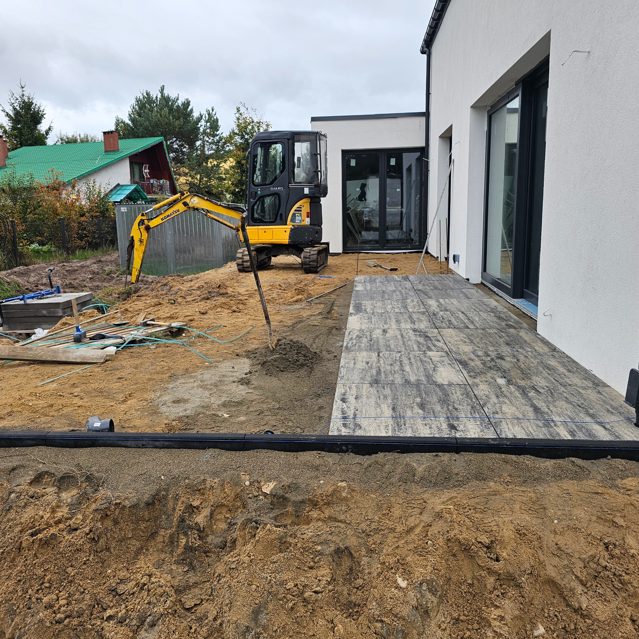 Układanie kostki brukowej na tarasie domu jednorodzinnego. Żółta koparka w tle. Widoczne przygotowane podłoże i częściowo ułożona nawierzchnia z szarych płyt.