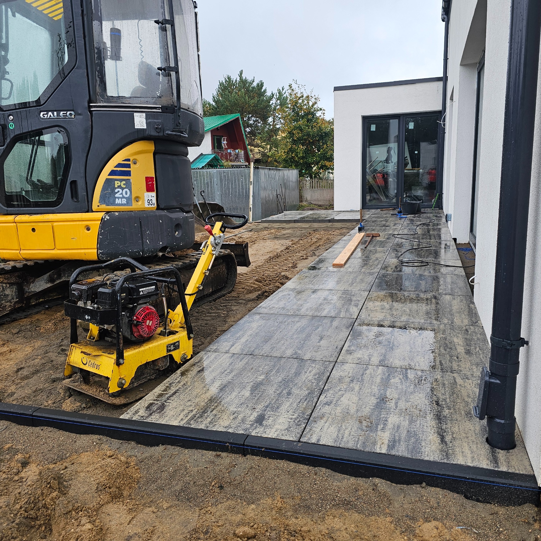 Układanie kostki brukowej na posesji z użyciem zagęszczarki i minikoparki. Widoczne świeżo ułożone płyty chodnikowe i przygotowane podłoże.