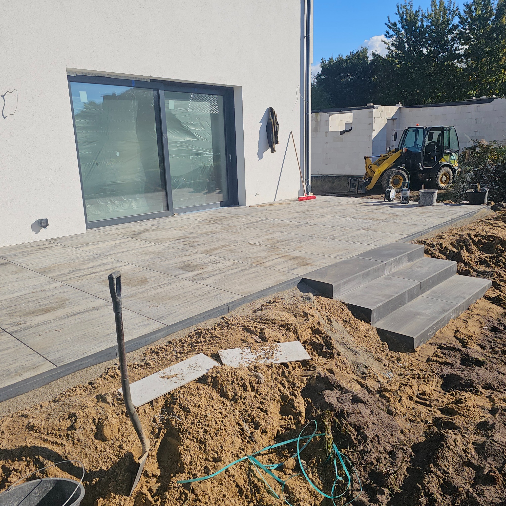 Układanie kostki brukowej na tarasie z widocznymi schodami, koparką w tle i narzędziami. Etap prac ziemnych, przygotowanie podłoża. Jasne płytki tarasowe.