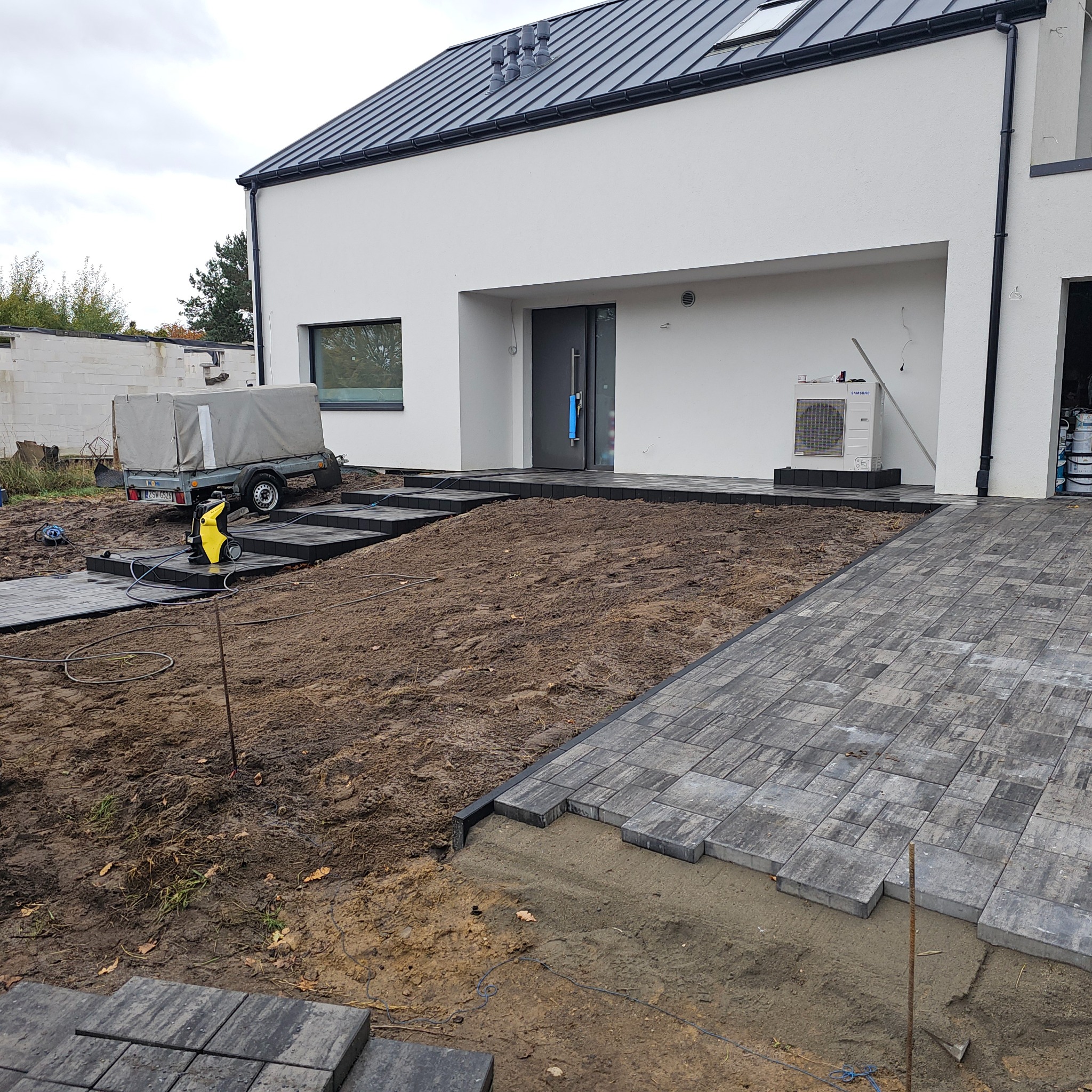 Układanie kostki brukowej na posesji nowoczesnego domu. Częściowo ułożona nawierzchnia z szarej kostki, widoczny sprzęt budowlany i przyczepka.