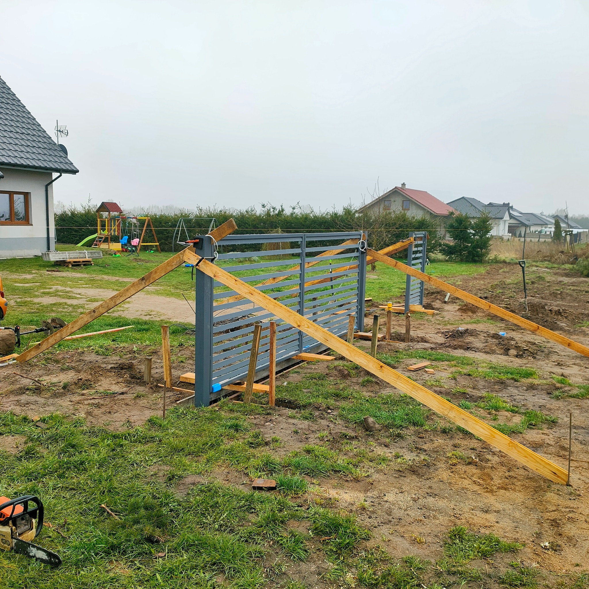 Montaż nowoczesnej, metalowej bramy wjazdowej na posesję. Widoczne drewniane podpory stabilizujące konstrukcję. W tle dom jednorodzinny i plac zabaw.