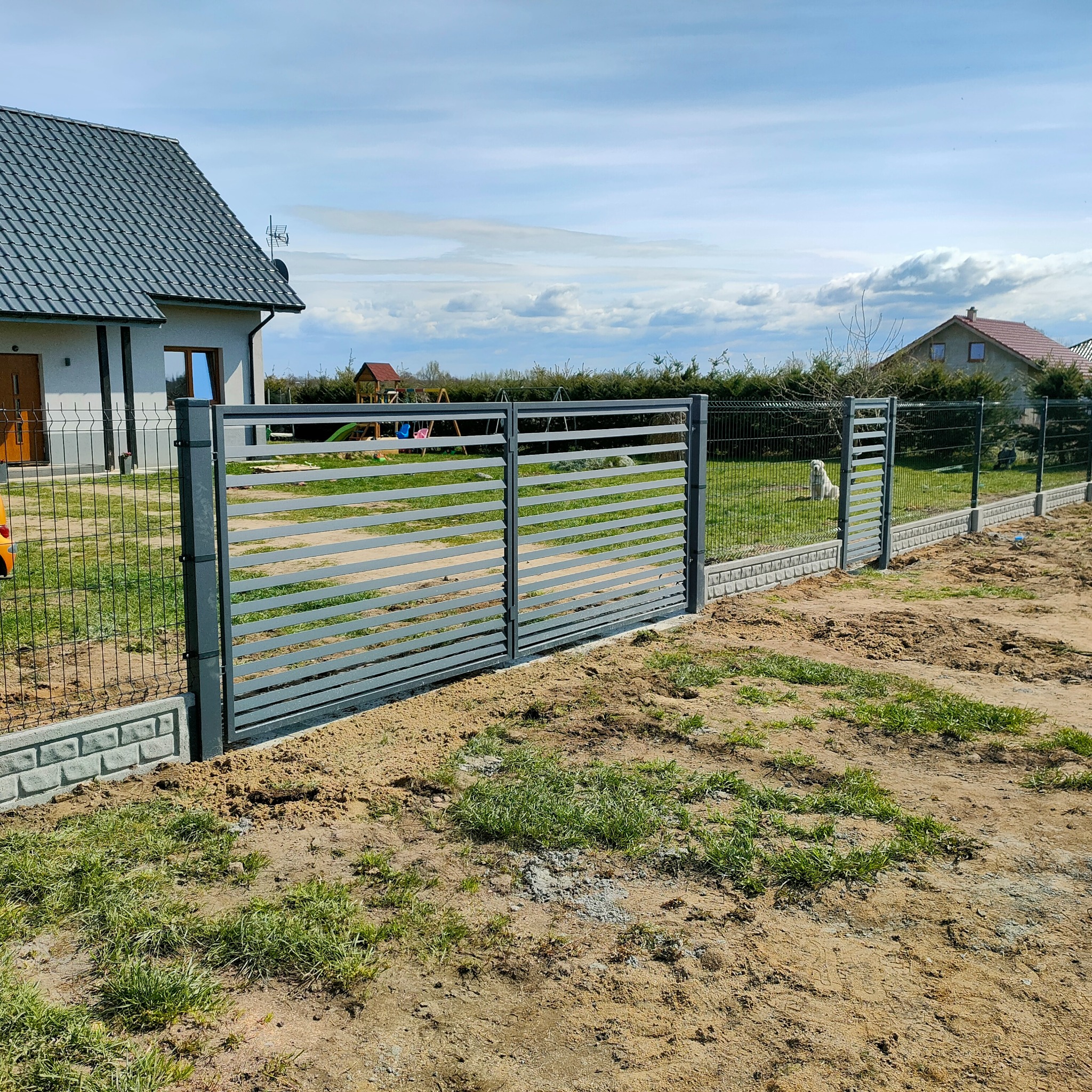 Nowoczesna, metalowa brama posesyjna z poziomymi szczeblami, osadzona na betonowej podmurówce. W tle dom z szarą dachówką i zielony trawnik.