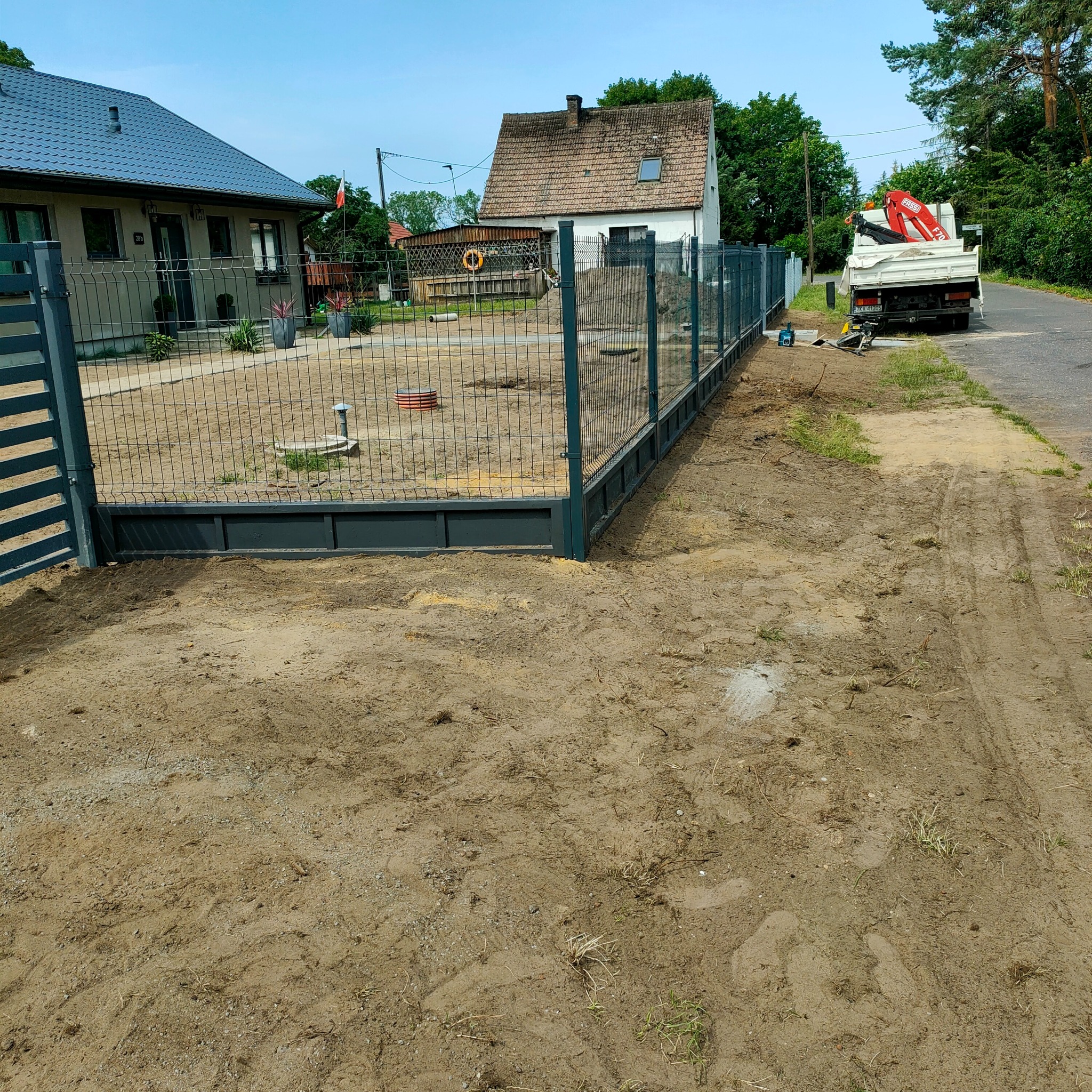 Nowoczesne ogrodzenie panelowe z metalową podmurówką, świeżo zamontowane na posesji. W tle domy i samochód ciężarowy z dźwigiem.