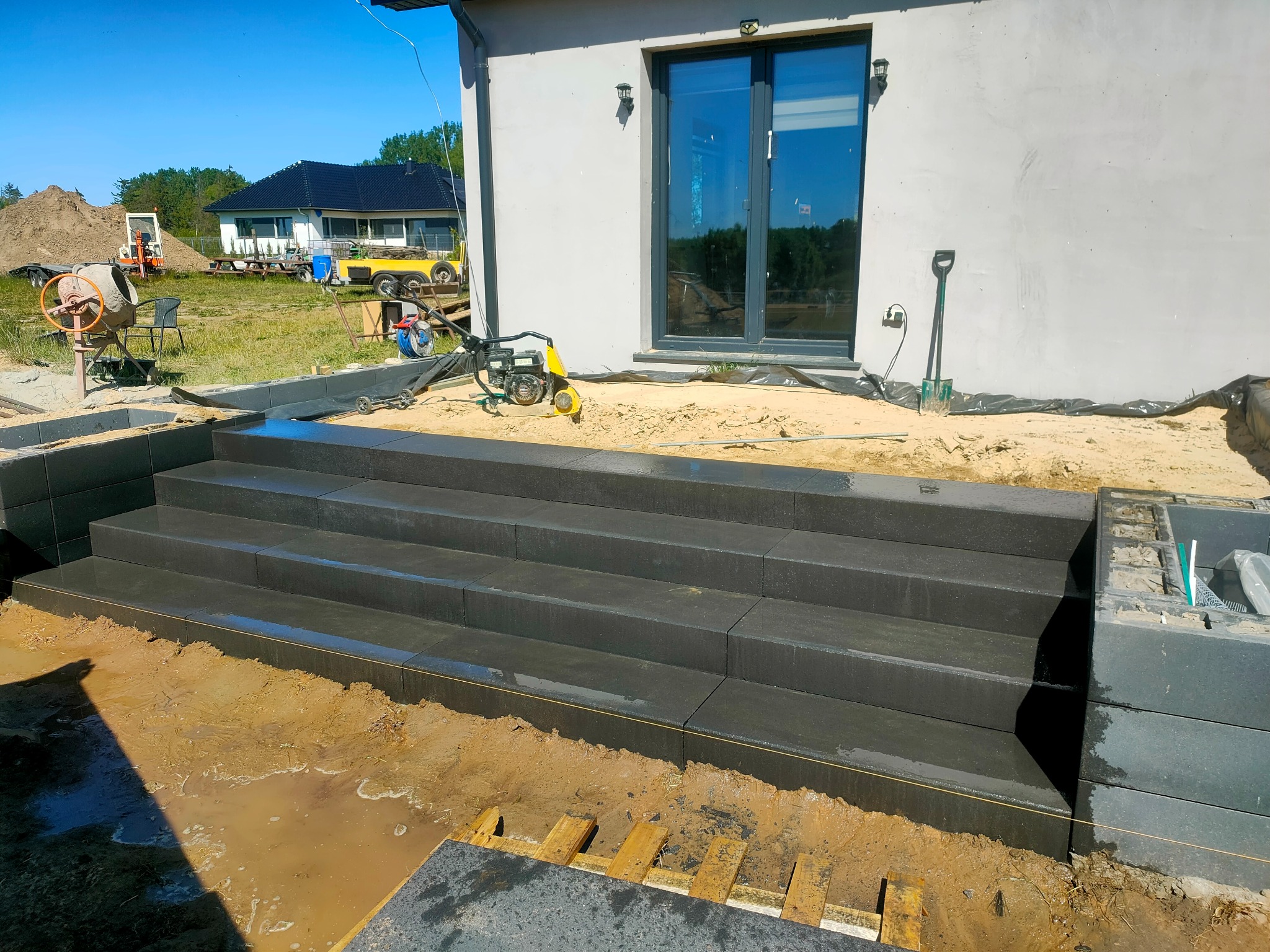 Nowe, ciemne schody wejściowe z kostki brukowej prowadzące do domu. Widoczny fragment elewacji, okno i narzędzia budowlane. Prace w toku, teren wokół rozkopany.