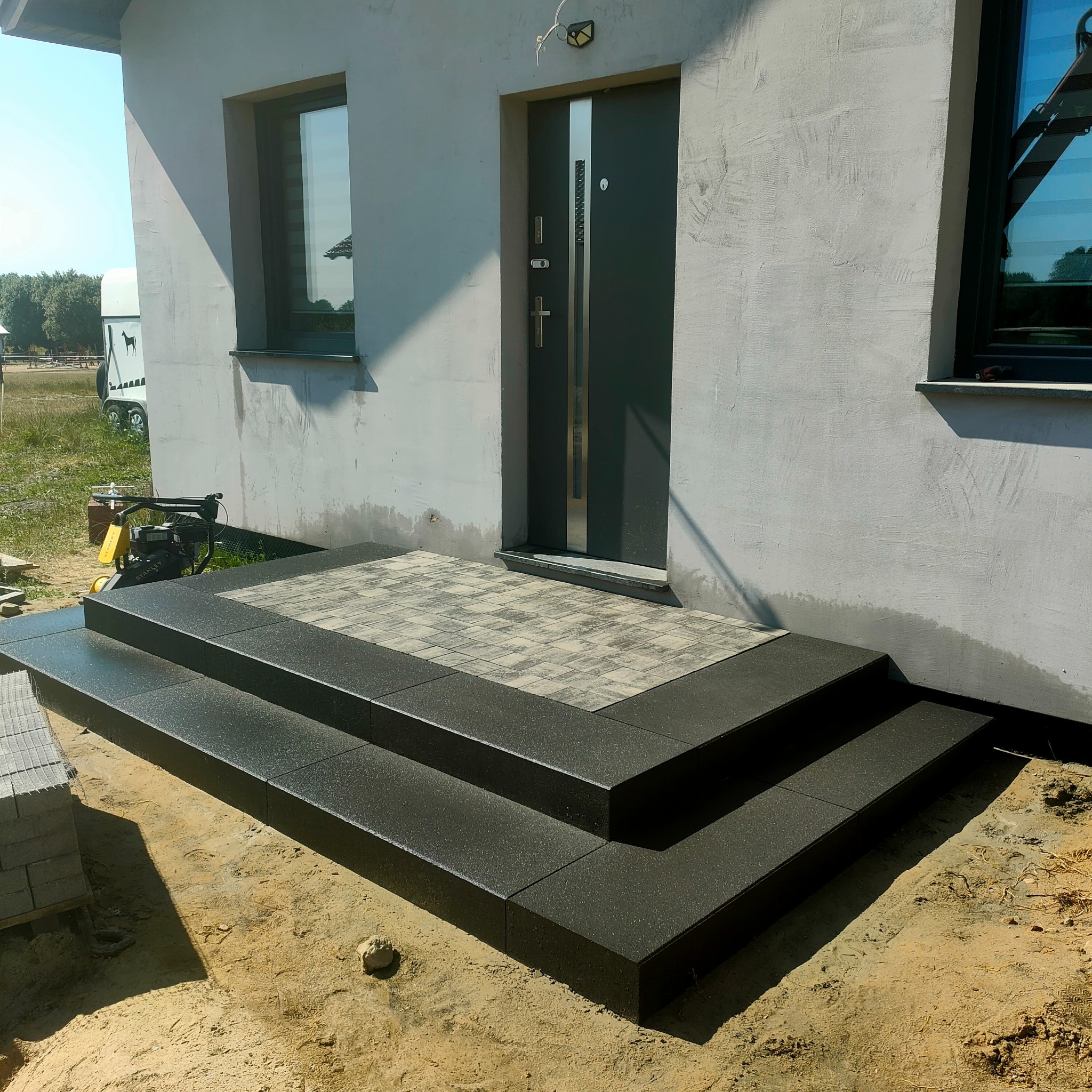 Nowe, dwustopniowe schody wejściowe z ciemnej kostki brukowej, prowadzące do szarych drzwi domu z oknami. Widoczna ubijarka Stanley i paleta kostki brukowej.