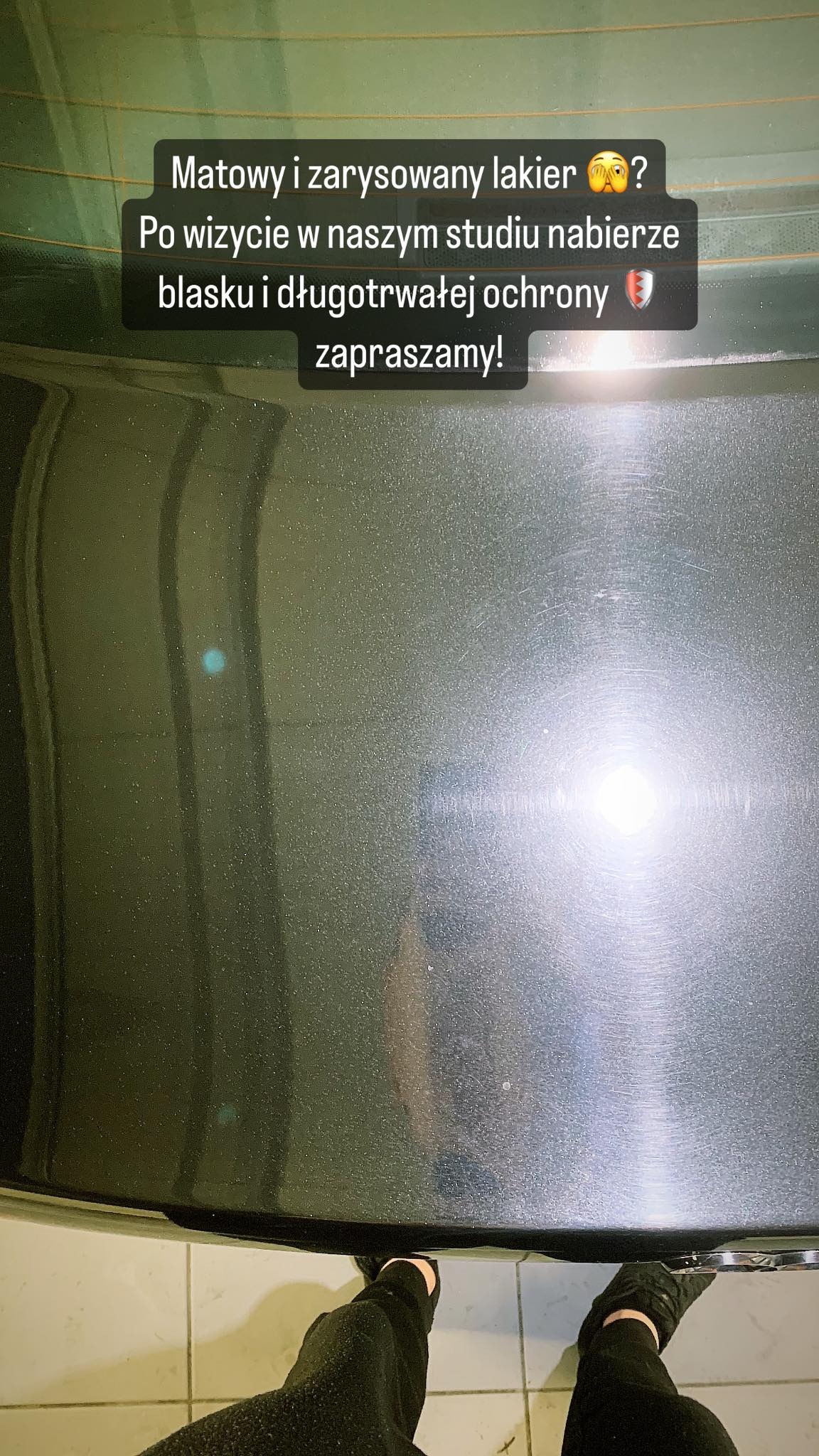 Fragment karoserii samochodu z widocznymi zarysowaniami i matowym lakierem, z refleksami światła, stojący na kafelkach, z fragmentem osoby w czarnych spodniach w dolnej części kadru.
