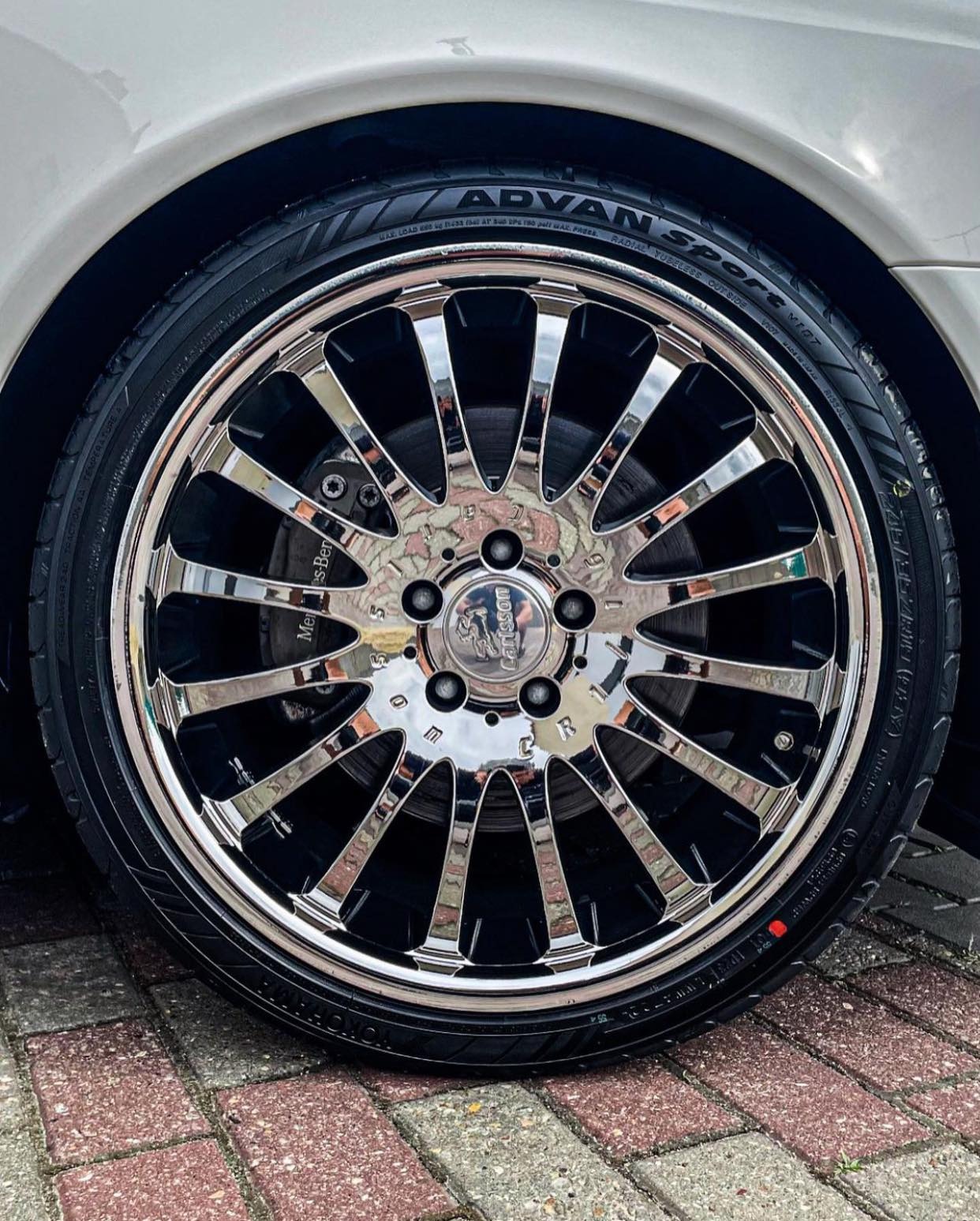Błyszczące, chromowane felgi Carlsson na oponie Advan Sport, widoczne zaciski hamulcowe Mercedes-Benz, ujęcie z bliska na tle kostki brukowej.
