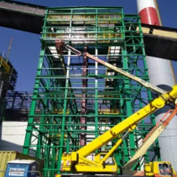 Montaż stalowej konstrukcji przemysłowej z użyciem podnośnika koszowego JLG Ultra Boom, widoczny komin i przenośna toaleta TOI-TOI.