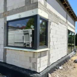 Montaż okna z czarną ramą w surowym budynku z betonu komórkowego; w szybie odbija się biały samochód dostawczy.