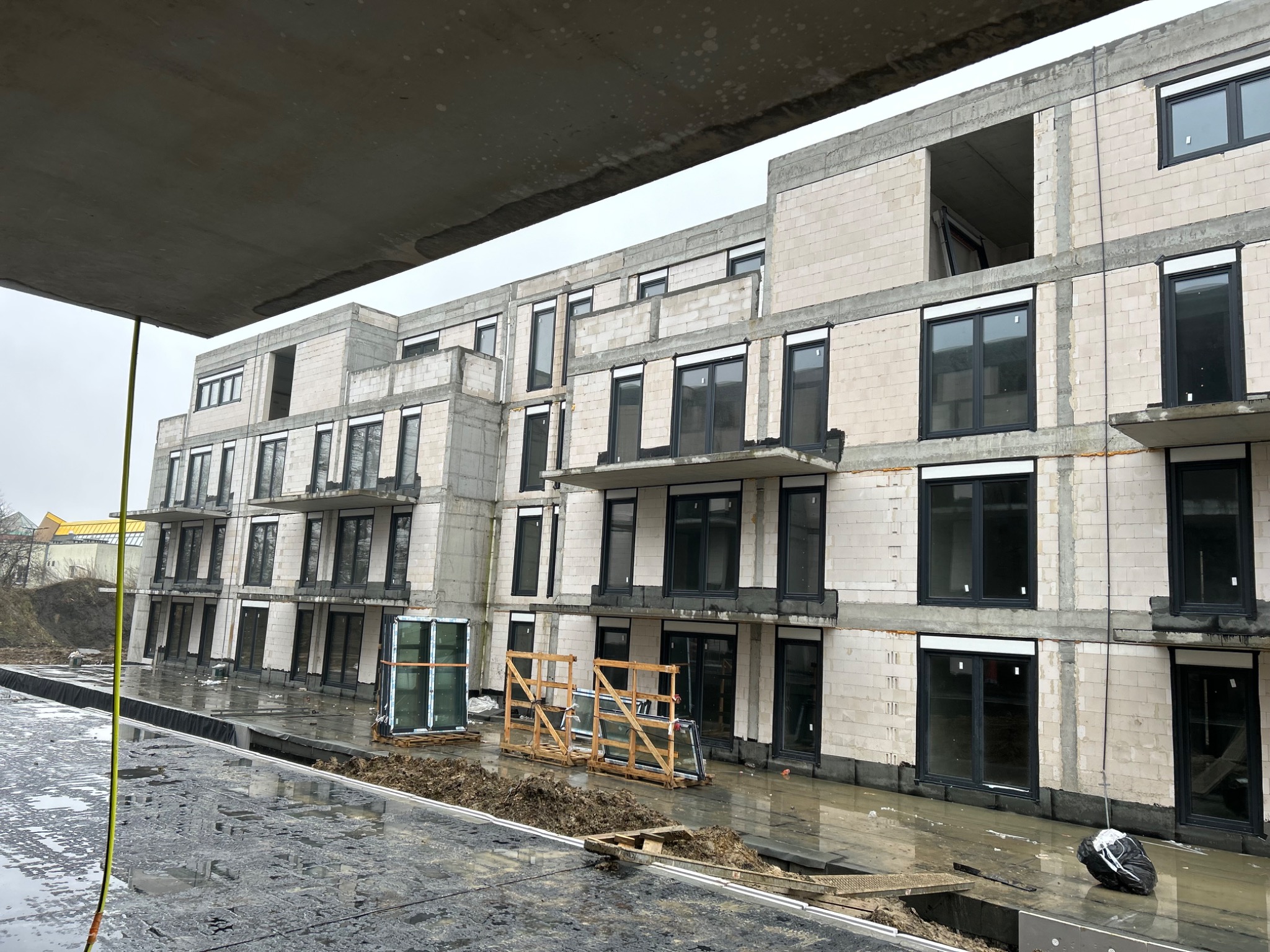 Budynek w trakcie montażu stolarki okiennej, widoczne okna z czarnymi ramami w surowym murze, palety z oknami czekające na montaż, mokry teren budowy, pochmurne niebo.