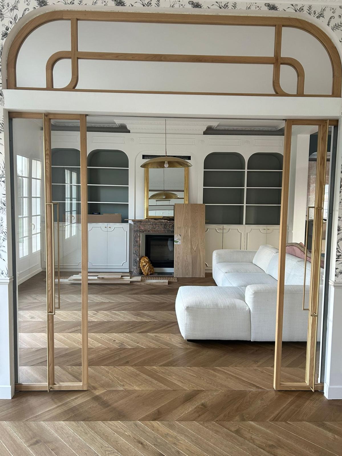 Salon z parkietem w jodełkę, przeszklonymi przesuwnymi drzwiami z drewnianą ramą, białym narożnikiem i kominkiem pod lustrem w złotej ramie, widok zza ozdobnego portalu z drewnianymi elementami.