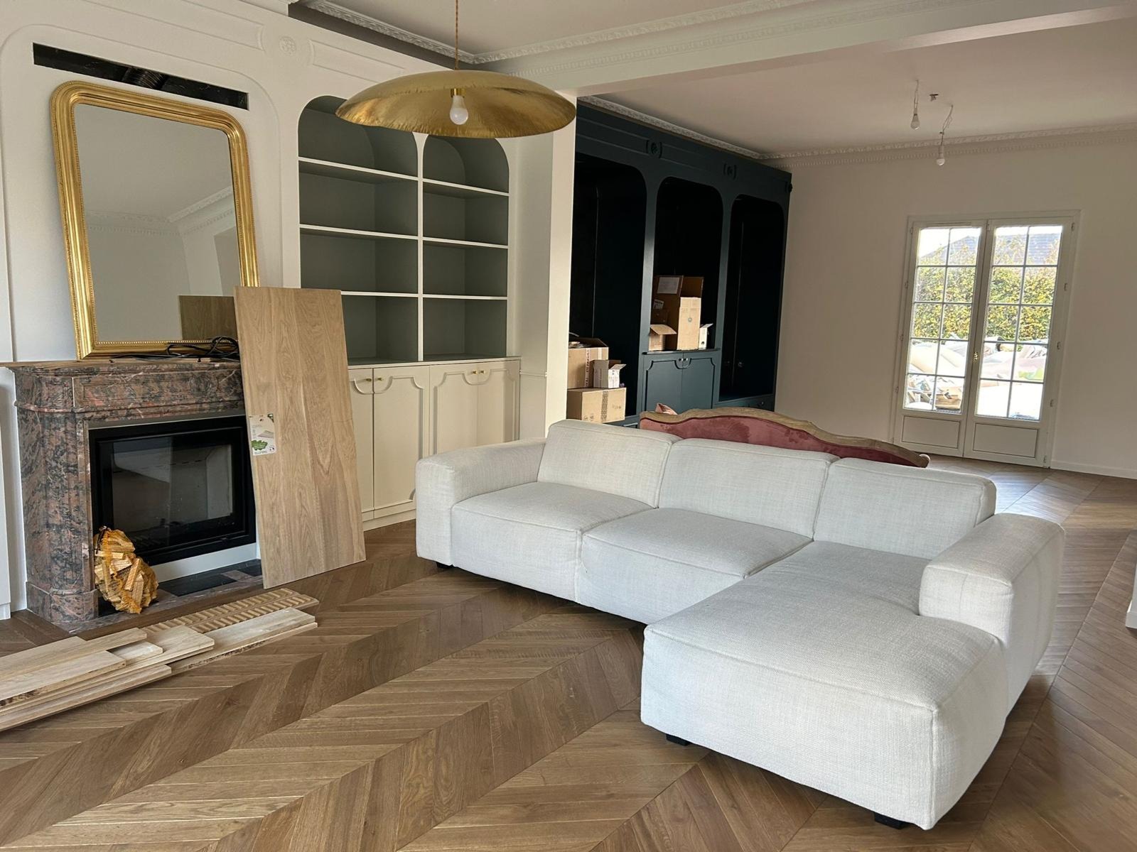 Salon w trakcie wykańczania: biała sofa modułowa, parkiet w jodełkę, marmurowy kominek z lustrem w złotej ramie, regał z półkami, kartony w tle.