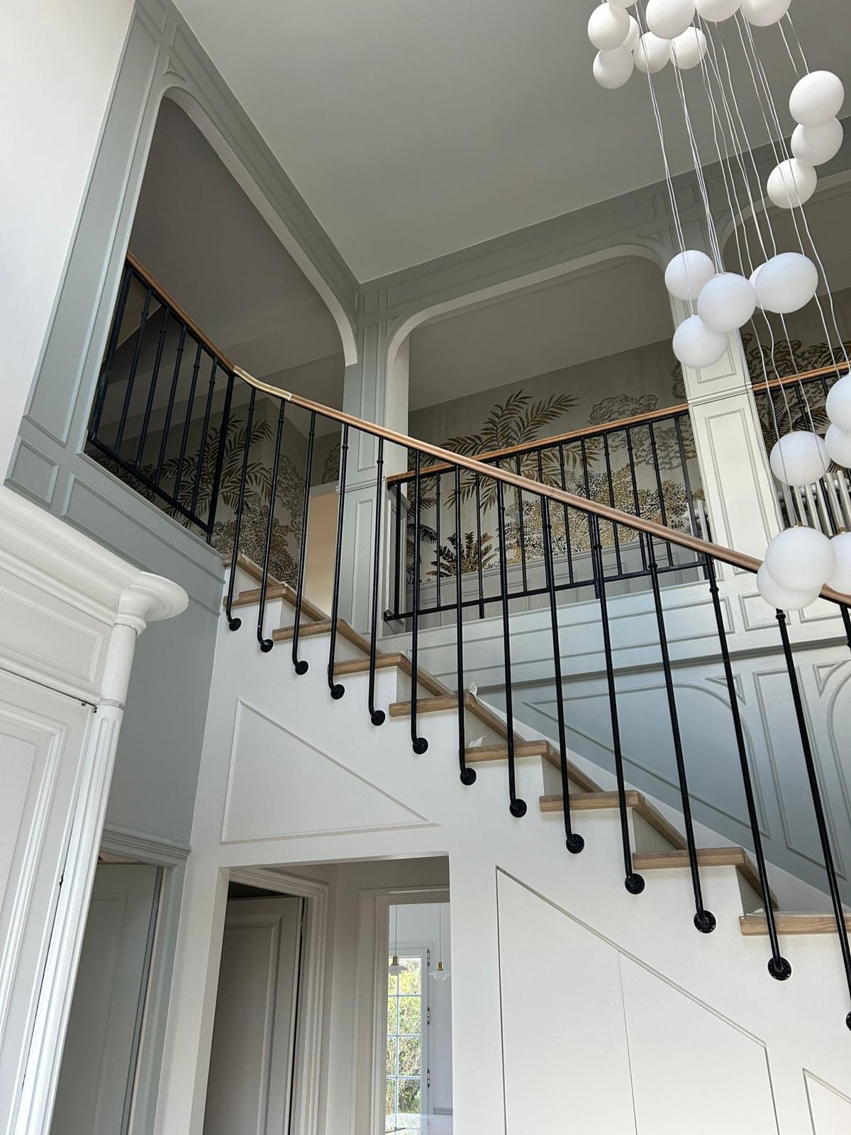 Klatka schodowa z drewnianymi stopniami, czarną balustradą i dekoracyjną tapetą z motywem tropikalnym, oświetlona nowoczesną lampą wiszącą z białych kul.