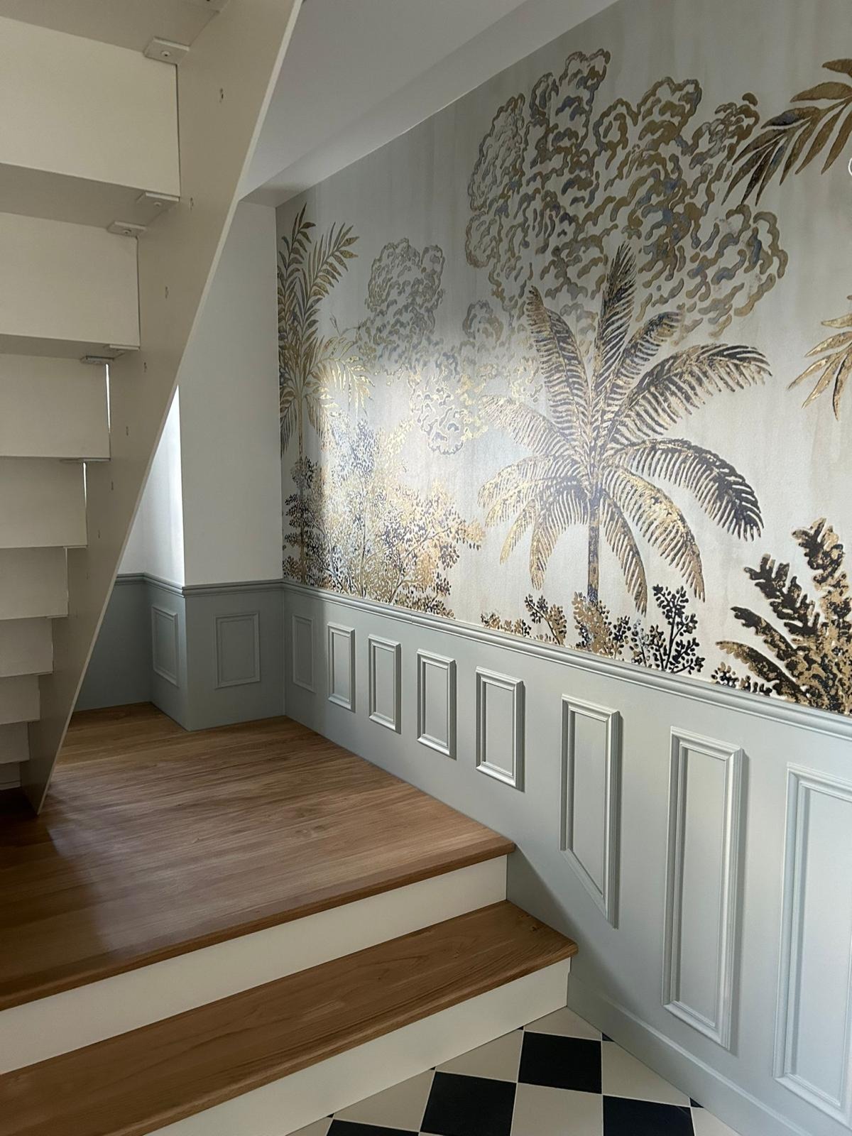 Wnętrze z tapetą w egzotyczne rośliny w odcieniach złota i szarości, listwy ścienne w kolorze gołębim, drewniane schody i czarno-biała szachownica na podłodze.