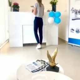 Recepcja w gabinecie fizjoterapii z białą ladą, dekoracjami z balonów i kwiatów, ulotki na stoliku kawowym z przodu, widok z perspektywy klienta.