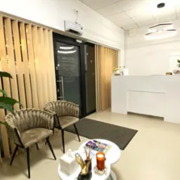 Jasne, przestronne wnętrze poczekalni z beżowymi krzesłami, białym biurkiem recepcyjnym, roślinami doniczkowymi i pionowymi żaluzjami, oświetlone nowoczesną lampą sufitową.