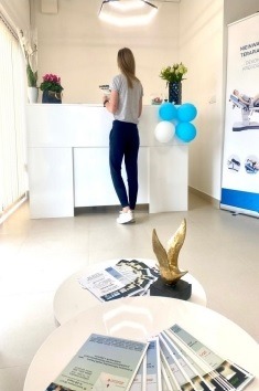 Recepcja w gabinecie fizjoterapii z białą ladą, dekoracjami z balonów i kwiatów, ulotki na stoliku kawowym z przodu, widok z perspektywy klienta.