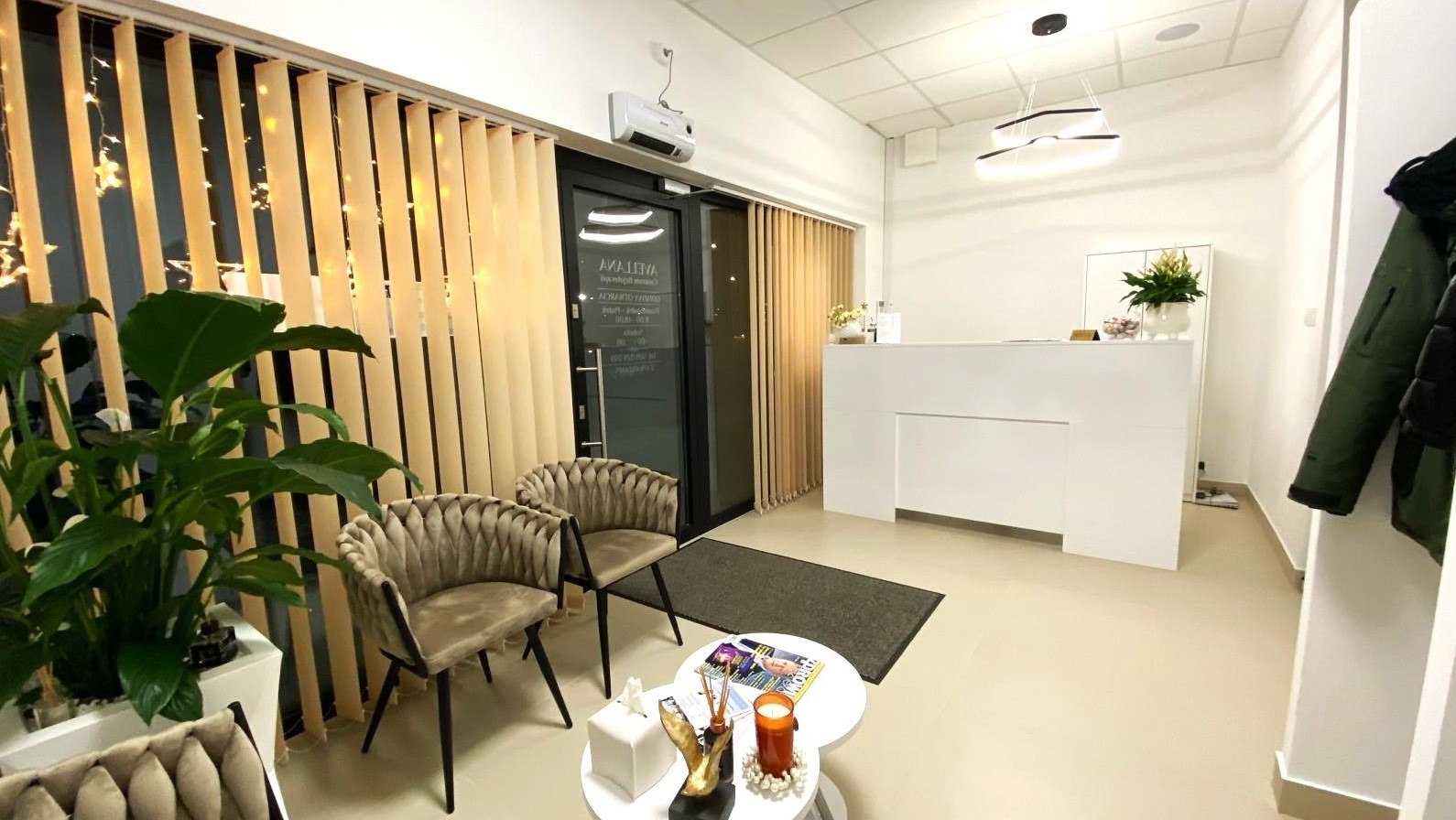 Jasne, przestronne wnętrze poczekalni z beżowymi krzesłami, białym biurkiem recepcyjnym, roślinami doniczkowymi i pionowymi żaluzjami, oświetlone nowoczesną lampą sufitową.