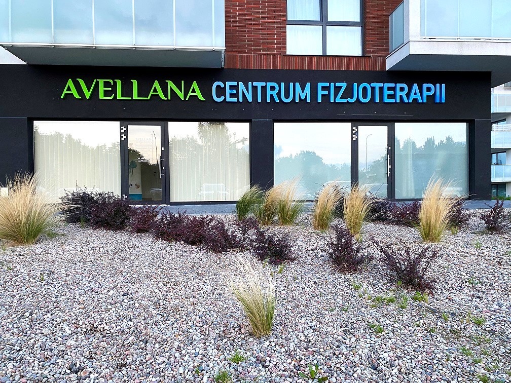 Nowoczesny budynek z napisem Avellana Centrum Fizjoterapii, otoczony ozdobnymi krzewami i kamieniami.