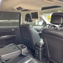 Taxi Rzeszow - Wnętrze czarnego samochodu z skórzanymi siedzeniami, widok z tyłu z rozkładanym podłokietnikiem i uchwytami na napoje, ekran zamontowany na suficie, widok na parking przez przyciemnione okno.