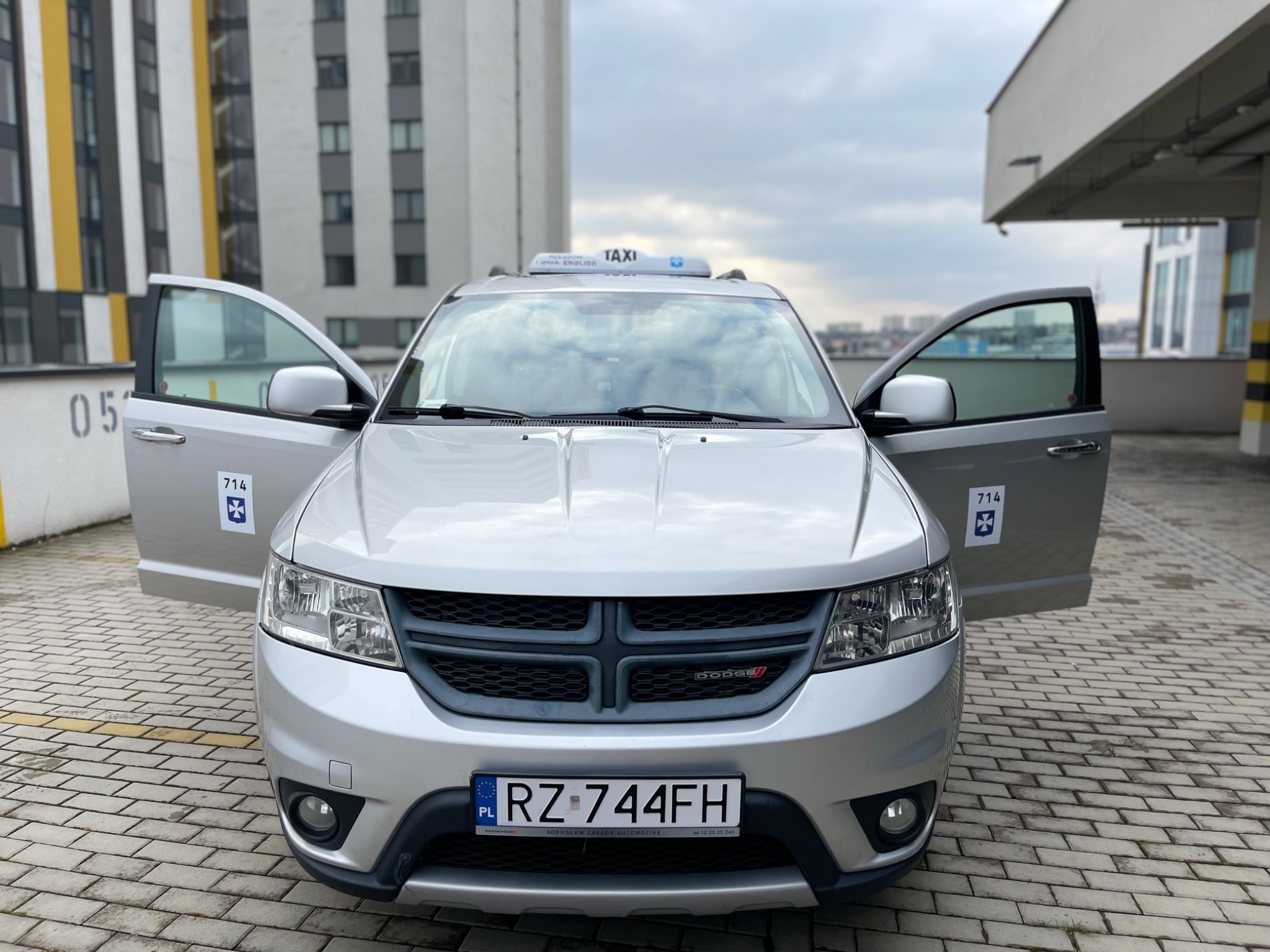 Srebrny Dodge Journey Taxi z otwartymi drzwiami, widok z przodu na tle budynku i pochmurnego nieba, numer rejestracyjny RZ 744FH.