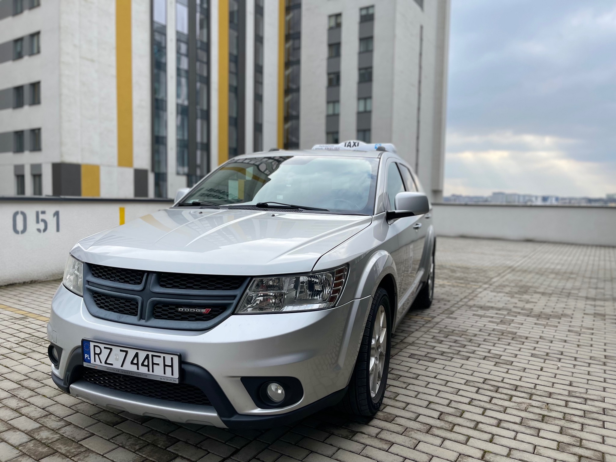 Srebrny samochód typu SUV z napisem TAXI na dachu, zaparkowany na brukowanym parkingu z budynkiem w tle.